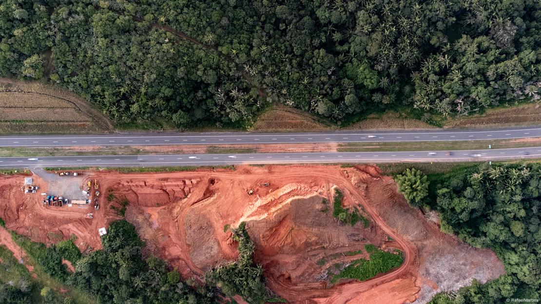 Do alto, imagem mostra duas vias de estrada asfaltada que divide paisagem. Acima da estrada, floresta; abaixo, um barranco de terra com área de estacionamento para caminhões.