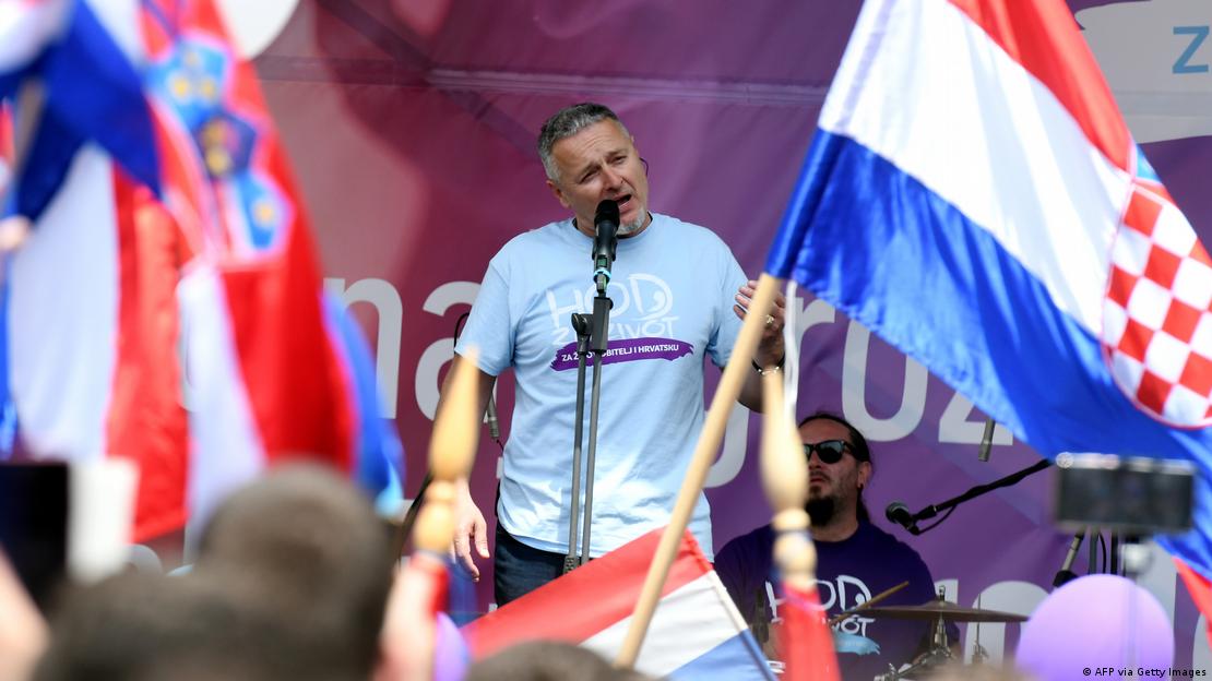 Thompson na bini ispred mikrofona s plavom majicom na kojoj piše "Hod za život" i hrvatskim zastavama uokolo