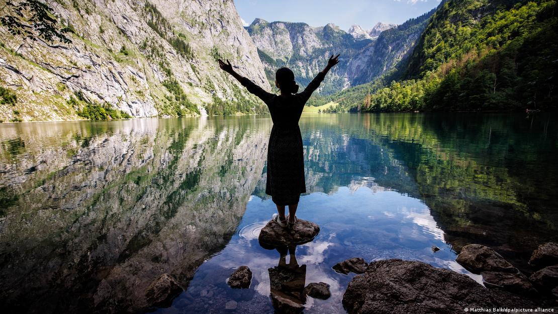 Eine junge Frau steht auf einem Stein im Wasser und blickt auf das umliegende Bergpanorama im Nationalpark Berchtesgaden Eine junge Frau steht auf einem Stein im Wasser und blickt auf das umliegende Bergpanorama im Nationalpark Berchtesgaden