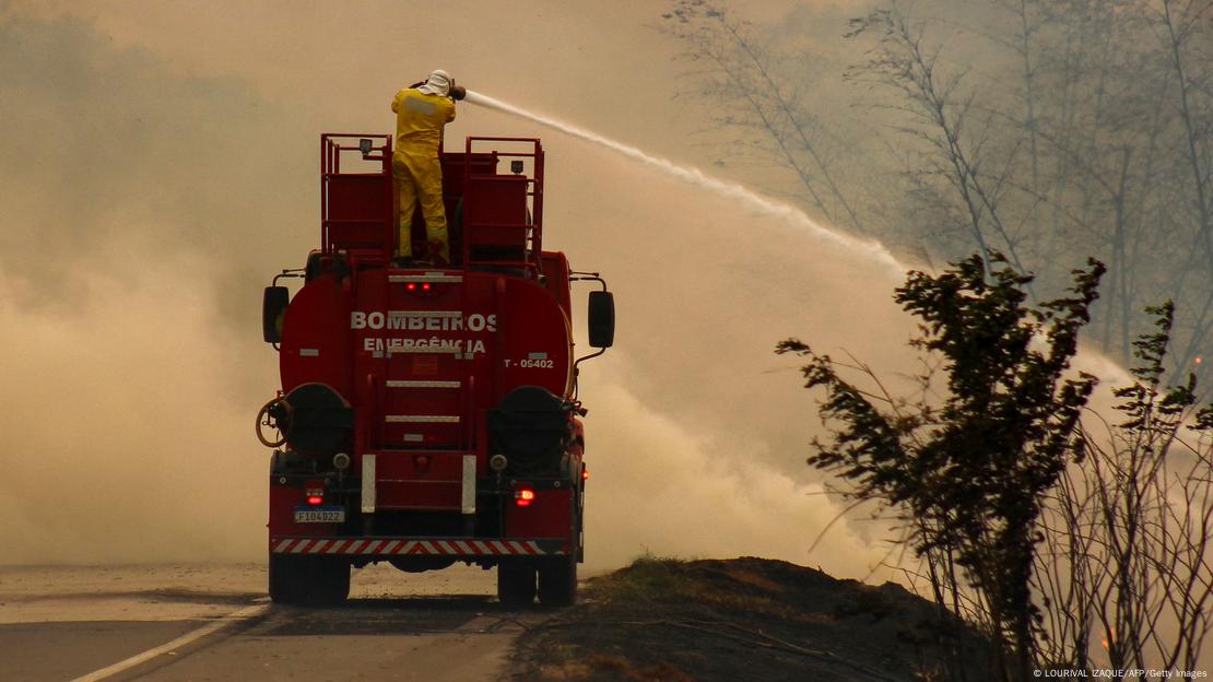 Homem vestido de amarelo sobre caminhão dos bombeiros direciona jato de água sobre incêndio na mata