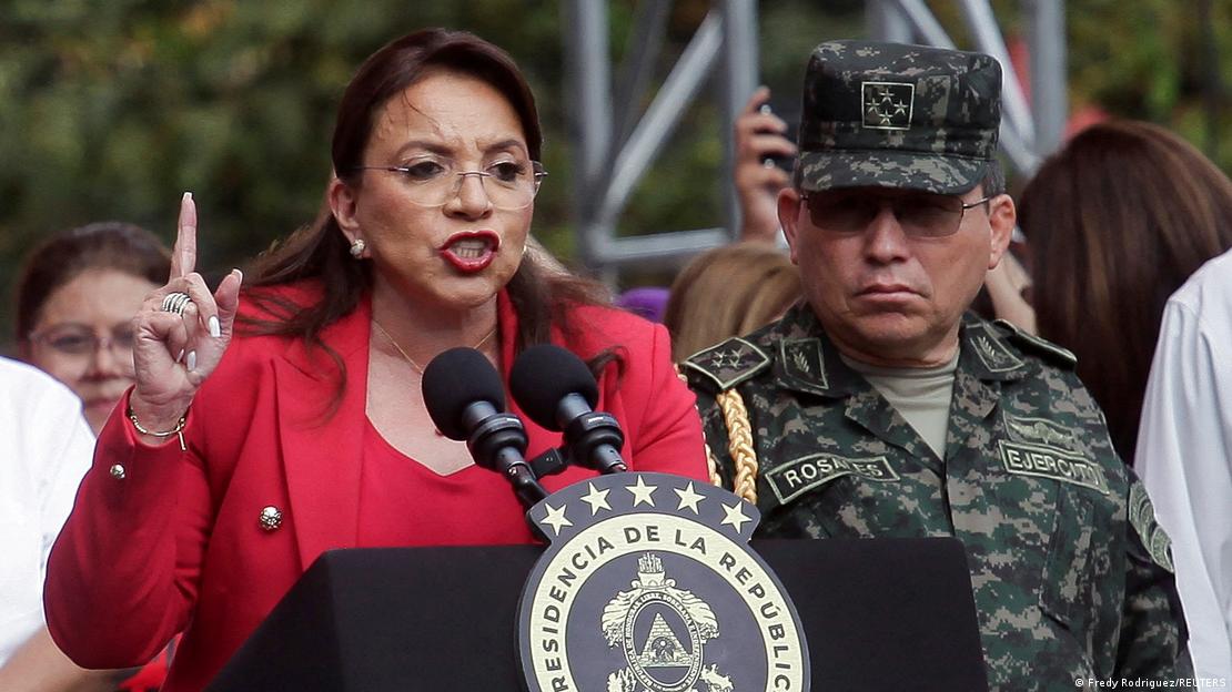 La presidenta de Honduras Xiomara Castro, habla a un micrófono, con el índice derecho en alto y vestida rojo, junto a un militar.
