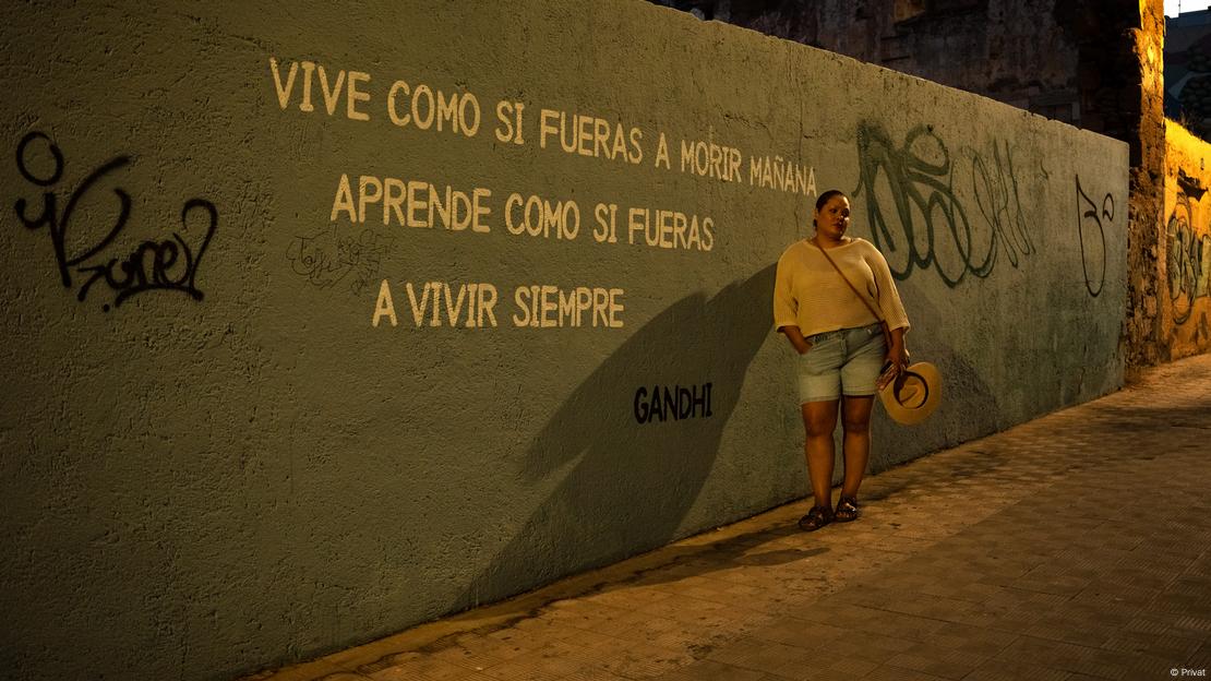 Kamila posa em frente a mural onde está escrito "Vive como si fueras a morir mañana/ Aprende como si fueras a vivir siempre", atribuída a Gandhi 