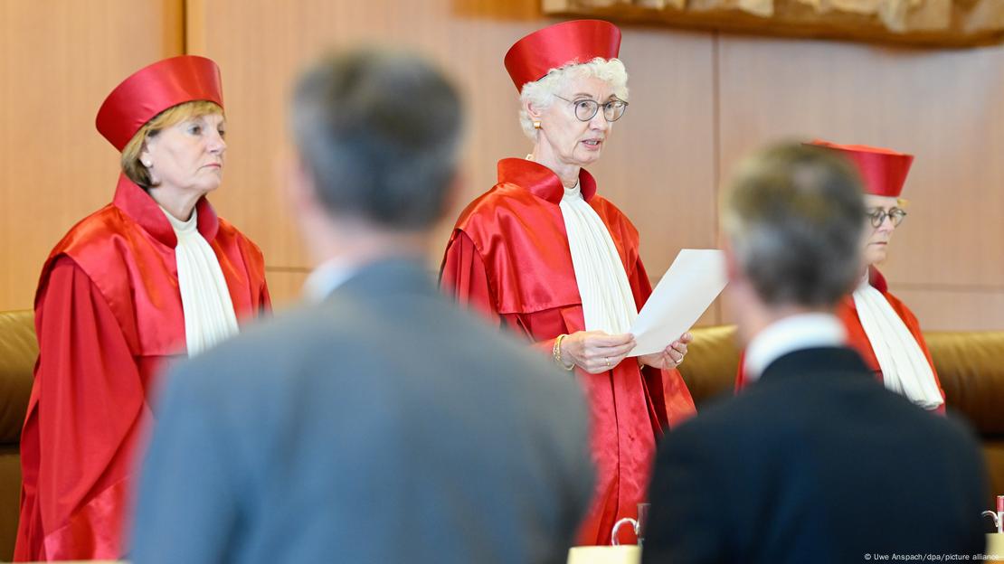 Drei Richterinnen in roten Roben und mit roter Kopfbedeckung stehen nebeneinander. Die Vorsitzende des Zweiten Senats, Doris König (M.), verkündet ein Urteil und hält dafür ein weißes Blatt Papier in der Hand.   