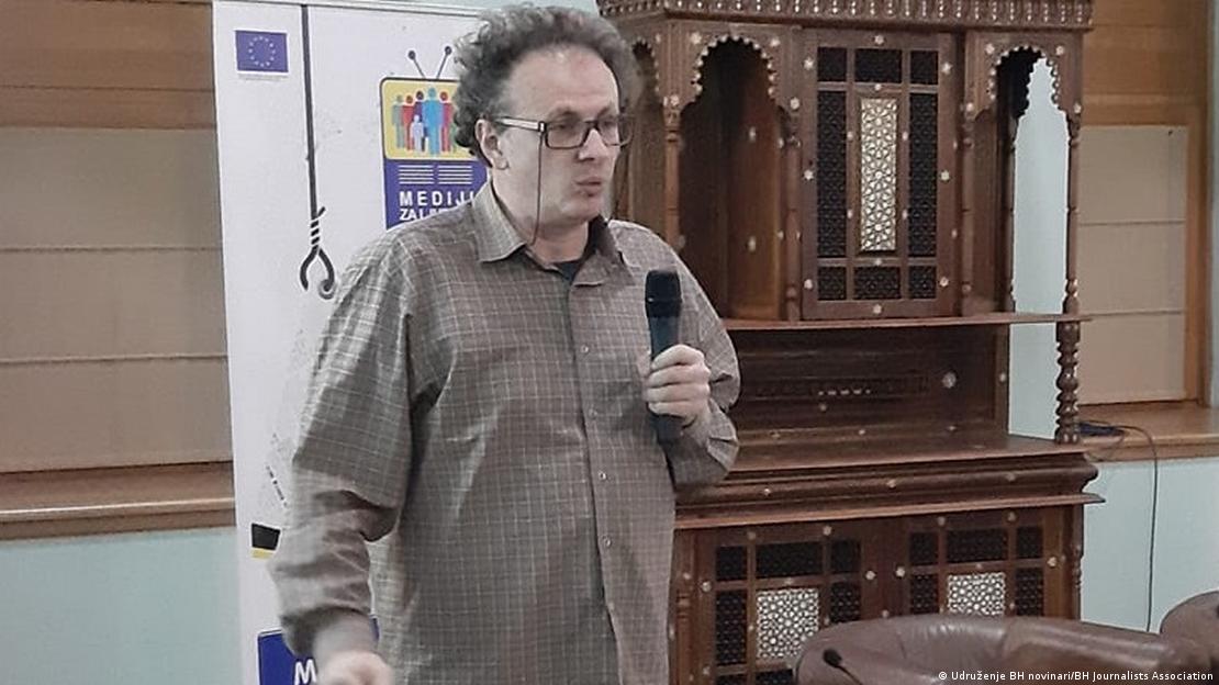 Muškarac čičkave kose i sa naočarama stoji i drži mikrofon