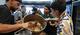 People stand around a metal table in a large kitchen and pour food from a pot into a flat shape