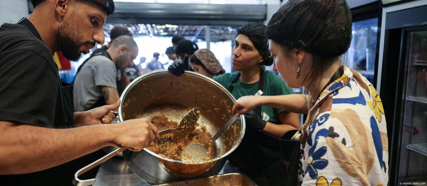 People stand around a metal table in a large kitchen and pour food from a pot into a flat shape