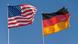 An American flag flies next to a German flag against a clear blue sky. An American flag flies next to a German flag against a clear blue sky.
