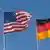 An American flag flies next to a German flag against a clear blue sky.