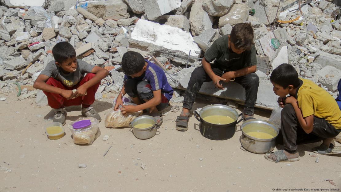 Menores palestinos junto a cazuelas con comida, sobre el suelo, sentados sobre restos de edificaciones destruidas.