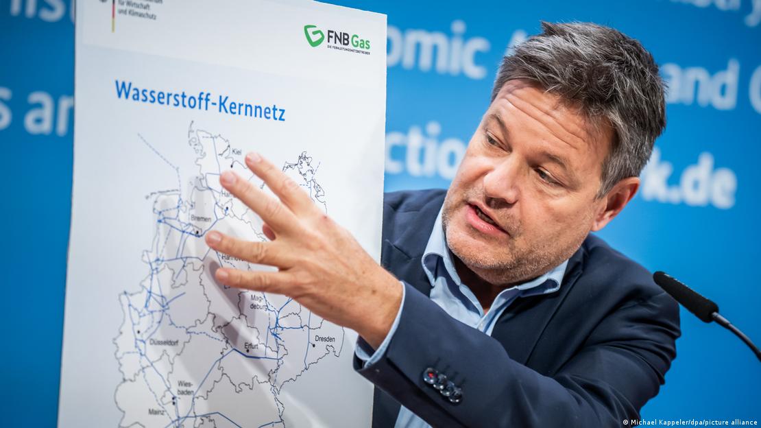 Deutschland Pläne für ein Wasserstoffkernnetz | Robert Habeck hält ein Schild hoch, auf dem der Umriss von Deutschland zu sehen ist. Innerhalb des Umrisses sind Leitungen eingezeichnet. Darüber steht: Wasserstoff-Kernnetz. Deutschland Pläne für ein Wasserstoffkernnetz | Robert Habeck hält ein Schild hoch, auf dem der Umriss von Deutschland zu sehen ist. Innerhalb des Umrisses sind Leitungen eingezeichnet. Darüber steht: Wasserstoff-Kernnetz.