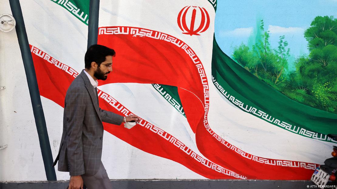 Un hombre vestido con saco sostiene un maletín en una mano y un tapabocas en la otra mientras pasa frente a un mural con la bandera iraní en Teherán.