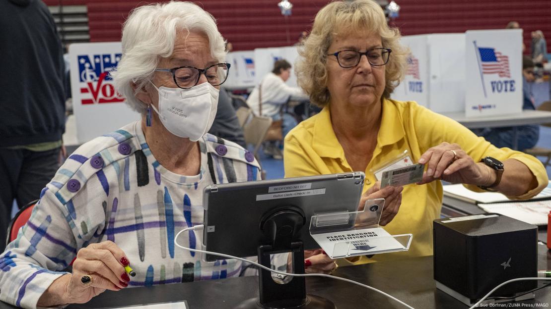 Zwei Frauen, eine von ihnen mit FFP2-Maske, begutachten einen Ausweis und ein Tablet. Im Hintergrund sind Wahlkabinen aufgestellt.