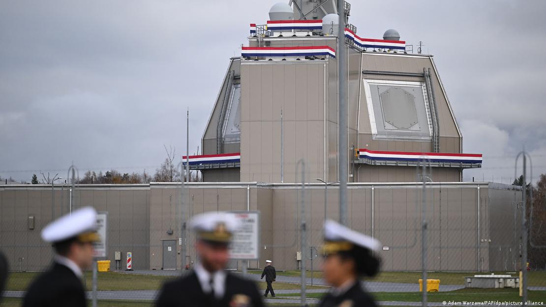 Ein graues Gebäude, das mit Fahnen mit rot-weiß-blauem Streifen geschmückt ist, hinter einet hohen grauen Mauer. Im Vordergrund sind unscharf drei Uniformierte zu sehen 