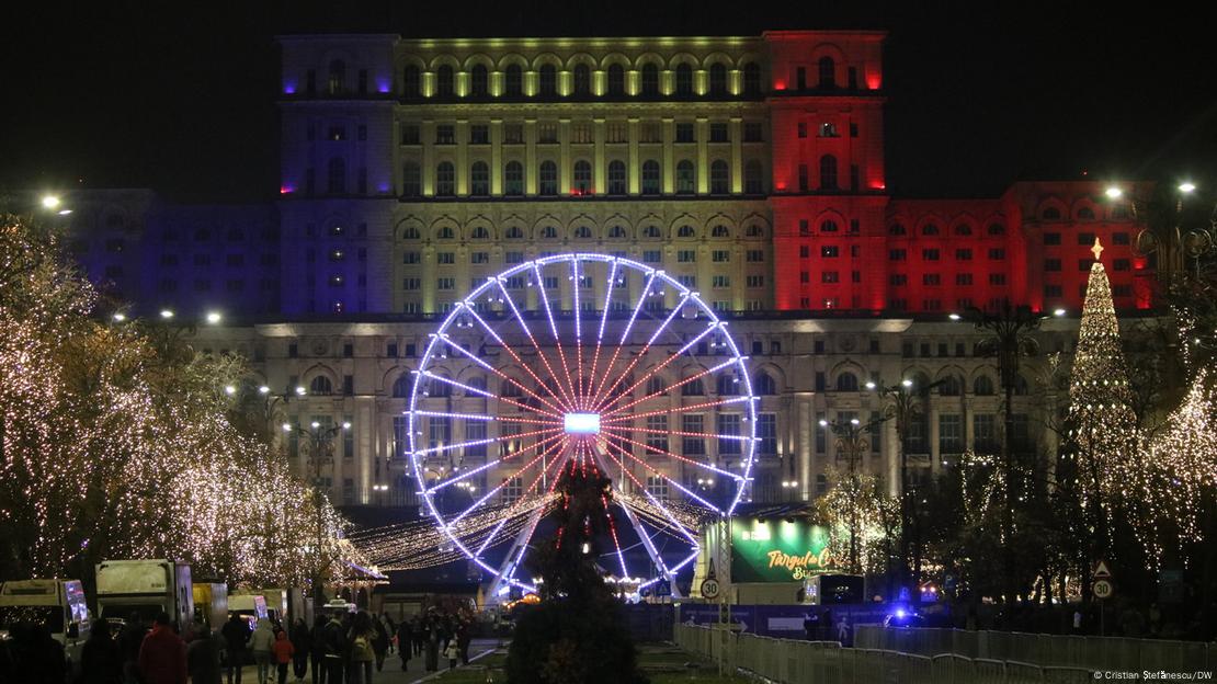 Ein erleuchtetes Riesenrad, dahinter ein sehr großes Gebäude, links und rechts beleuchtete Bäume