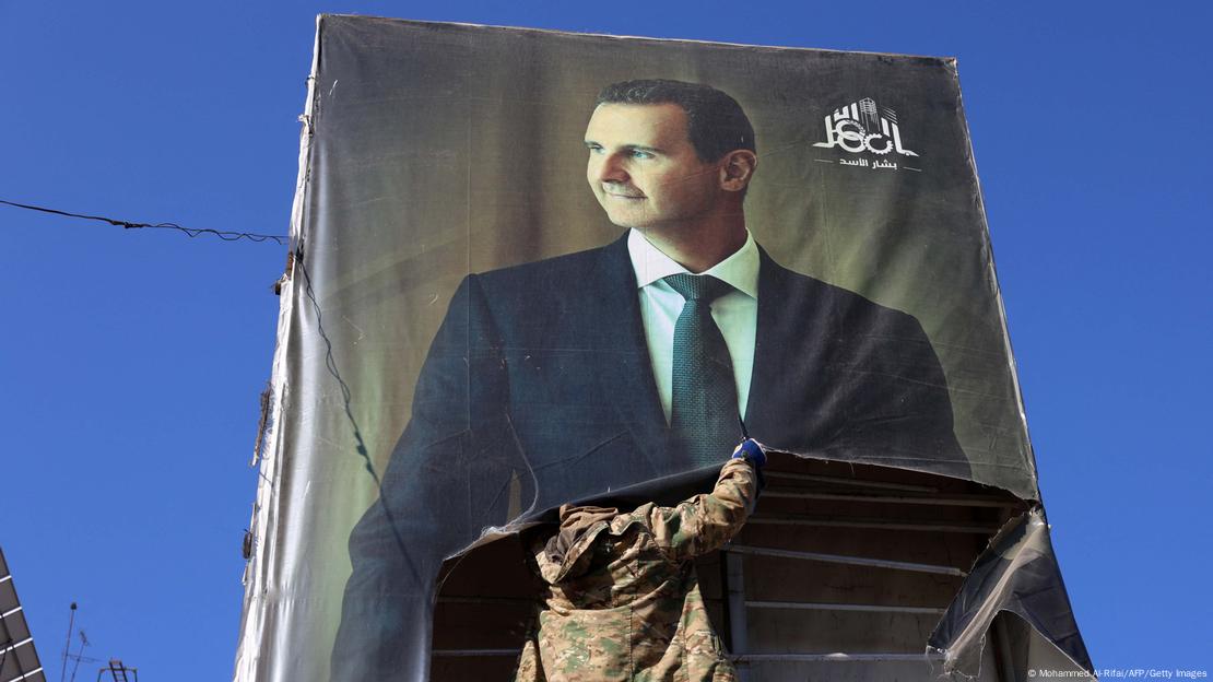 Um grande cartaz com a imagem do ex-ditador sírio Bashar al-Assad é retirado por um rebelde que veste roupas militares. Um grande cartaz com a imagem do ex-ditador sírio Bashar al-Assad é retirado por um rebelde que veste roupas militares.