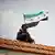 A Syrian with a gun standing on a roof. A Syrian flag flies above him