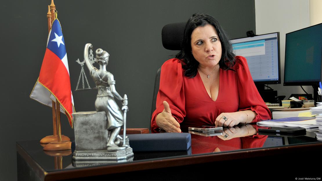 Trinidad Steinert sentada em mesa, sobre a qual há uma bandeira do Chile e uma pequena estátua que representa a Justiça Trinidad Steinert sentada em mesa, sobre a qual há uma bandeira do Chile e uma pequena estátua que representa a Justiça