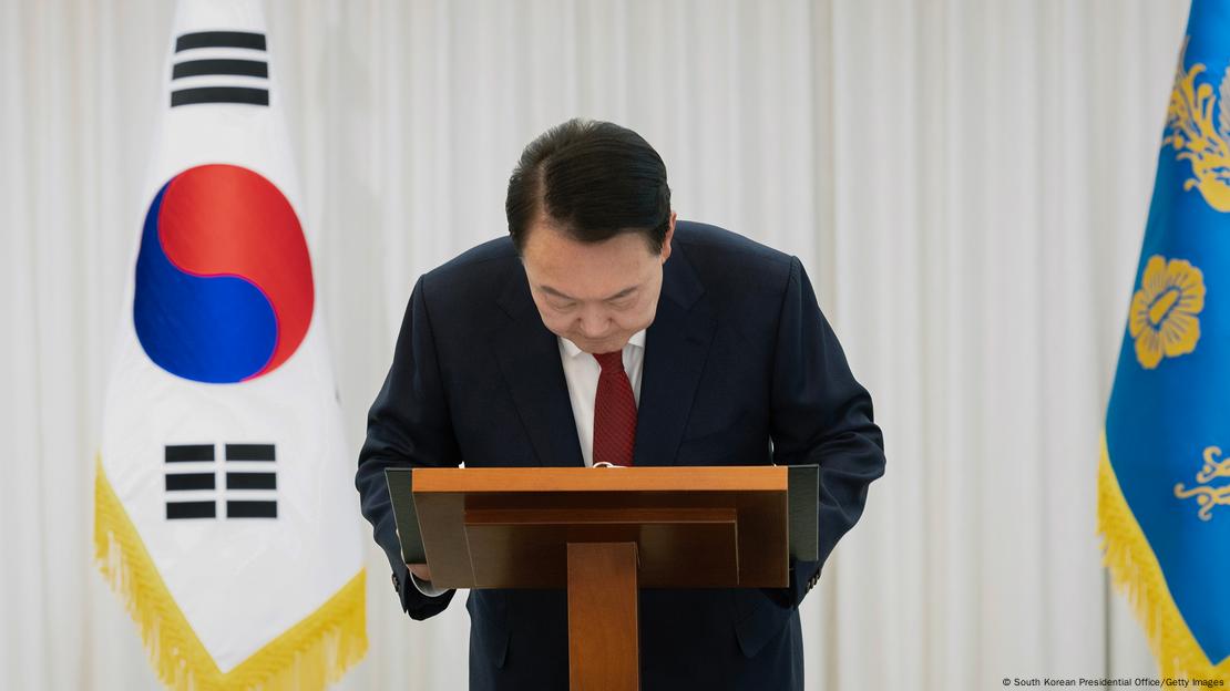 Südkoreas suspendierter Präsident Yoon Suk Yeol an einem Sprecherpult, er beugt den Kopf und Oberkörper nach vorne, im Hintergrund die Landesflagge