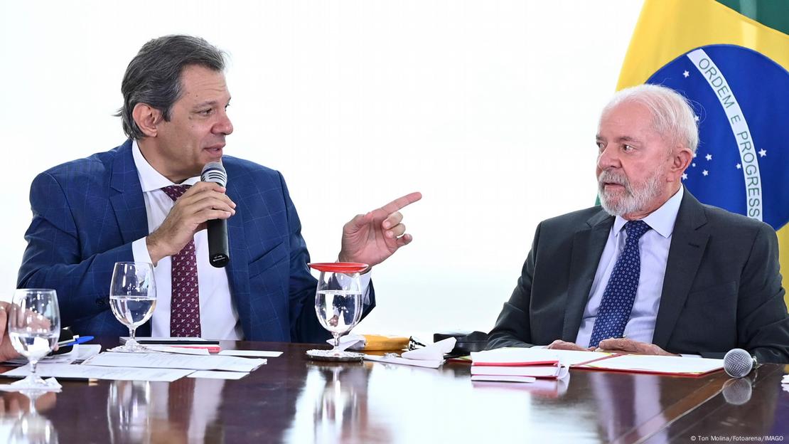 Haddad e Lula lado a lado sentados numa mesa de reunião. Haddad aponta para Lula.