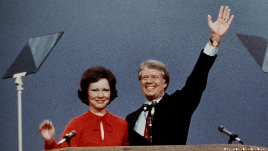 Carter duke përshëndetur me dorën lart, dhe bashkëshortja veshur me xhaketë të kuqe duke buzëqeshur