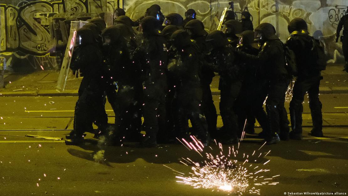 Grupo de policiais se protegendo com escudos em uma rua, enquanto fogo de artifício explode ao lado