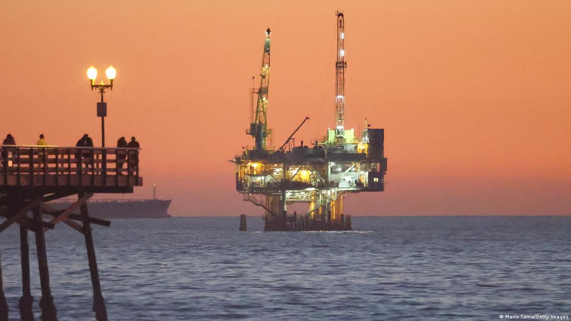 Plataforma de petróleo iluminada durante um entardecer observada por pessoas em um pier nos EUA
