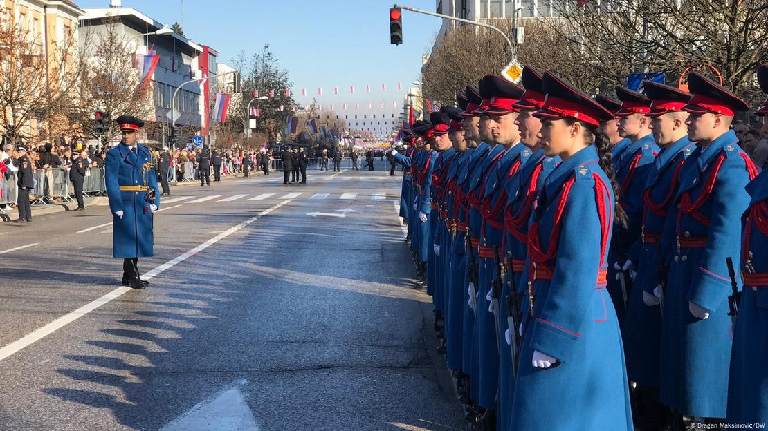 Na slici se vidi postrojavanje počasne garde u plavim uniformama s crvenim detaljima i kapama. Garde su poredane uz ulicu, dok je u pozadini vidljiva publika, zgrade ukrašene zastavama i svijetli zimski dan.