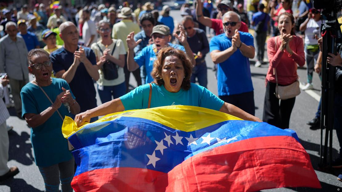 Protesta en Caracas contra Nicolás Maduro antes de su tercera asunción del cargo como presidente: Una mujer con una bandera de Venezuela, y un grupo de personas detrás. Protesta en Caracas contra Nicolás Maduro antes de su tercera asunción del cargo como presidente: Una mujer con una bandera de Venezuela, y un grupo de personas detrás.