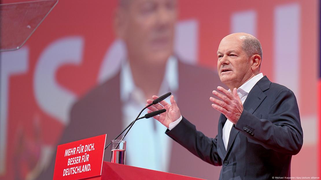 Bundeskanzler Olaf Scholz (SPD) spricht beim außerordentlichen SPD-Bundesparteitag zu den Delegierten. Er trägt einen dunklen Anzug und ein weißes Hemd ohne Krawatte. Hinter ihm ist eine Videoanzeige zu sehen, auf der er ebenfalls zu sehen ist. An seinem Rednerpult steh sein Wahlkampfspruch: "Mehr für dich. Besser für Deutschland."