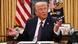 O presidente dos Estados Unidos, Donald Trump, segura uma caneta após assinar um decreto no Salão Oval da Casa Branca. Ele veste terno e gravata escuros, e camisa branca. Ao fundo, está a bandeira dos Estados Unidos e fotos da sua família.