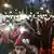 A woman raises a red-gloved hand in protest at a demonstration in support of teachers in Belgrade, Serbia, January 19, 2025. It is night time. In the background are many other protesters, some holding up signs; others holding up cell phones with the lights switched on