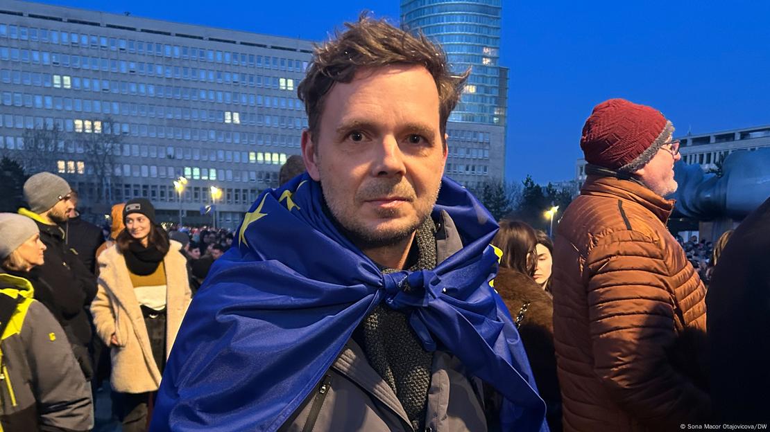 A man with a large EU flag tied around his shoulders can be seen in the middle of a crowd as darkness falls. He is looking straight into the camera. Bratislava, Slovakia, January 24, 2025