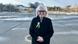 A woman (Lucia Stasselova) dressed in a black overcoat and with a blue-and-yellow badge and ribbons on her lapel stands in an open space in front of a large metal fountain and smiles into the camera, Freedom Square, Bratislava, Slovakia, January 24, 2025