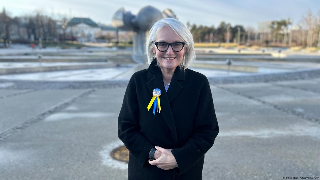 A woman (Lucia Stasselova) dressed in a black overcoat and with a blue-and-yellow badge and ribbons on her lapel stands in an open space in front of a large metal fountain and smiles into the camera, Freedom Square, Bratislava, Slovakia, January 24, 2025