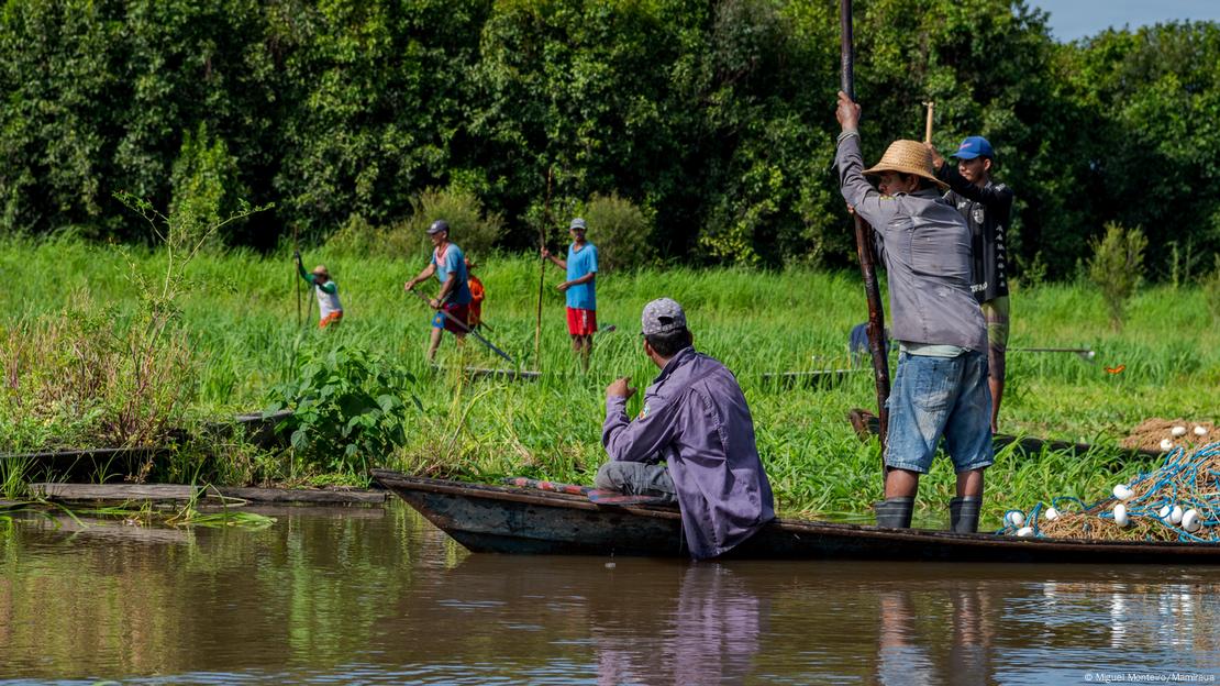 Homens dentro de canoa em rio, observados por outros homens em outra canoa, atrás de área com mato