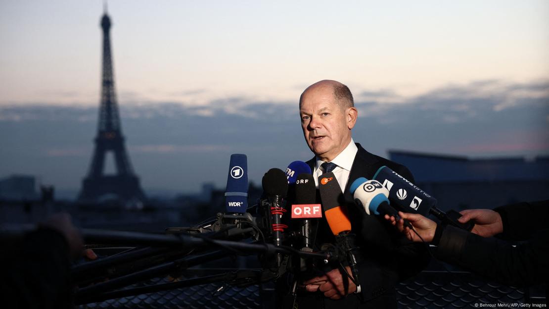 Olaf Scholz fala diante de microfones segurados por repórteres, com a Torre Eiffel ao fundo, ao entardecer