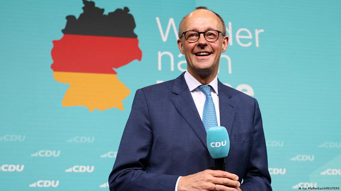 Bundestagswahl 2025. Friedrich Merz steht vor einer türkisfarbenen Wand, auf der das CDU-Logo mehrfach zu lesen ist, darüber die Umrisse Deutschlands in den deutschen Nationalfarben Schwarz, Rot und Gold. Merz hält ein Mikrofon in der Hand und lacht. 