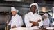 A Black man stands with his arms crossed in a kitchen. Behind him, kitchen workers cook. A Black man stands with his arms crossed in a kitchen. Behind him, kitchen workers cook.
