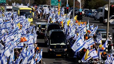 People hold Israeli flags as a funeral convoy of black limousines makes its way down a road in Ashkelon, Israel