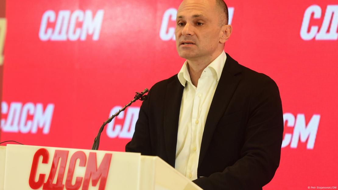 Маж во црно сако (Венко Филипче) стои пред микрофон на говорница, на која и зад која се гледа логото на СДСМ 