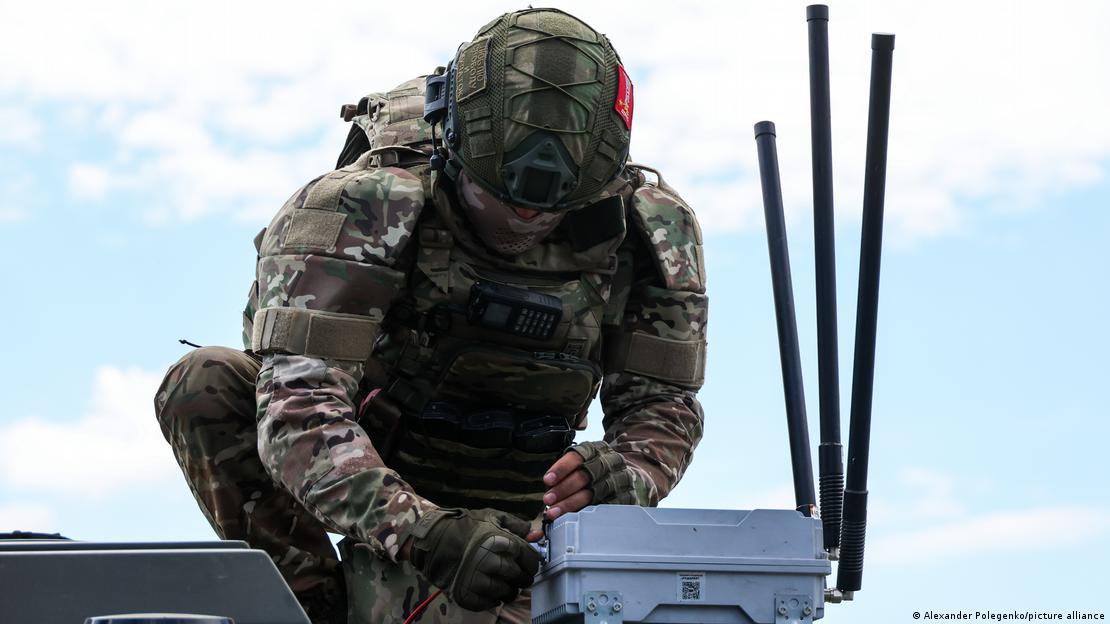 A soldier in camouflage uniform and helmet, photographed from below against a pale blue sky, crouches over a plastic box with three antennae sticking up