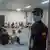 A security guard wearing a medical mask stands at the door with civilians sitting on the floor behind them