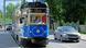 A blue tram on a street, a car running alongside it A blue tram on a street, a car running alongside it