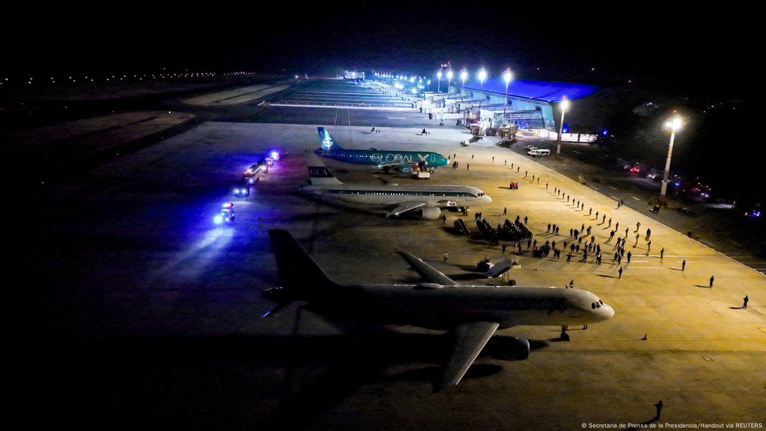 Varios aviones aparcados en hilera y personas andando por la pista de un aeropuerto. 