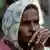 A woman in a headscarf with blurred face, hands clasped, greenery in background