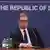 President Aleksandar Vucic looks serious as he sits at a table in front of a blue backdrop emblazoned with the words 'Republic of Serbia'. There are two bottles of water, a glass and a microphone in front of him