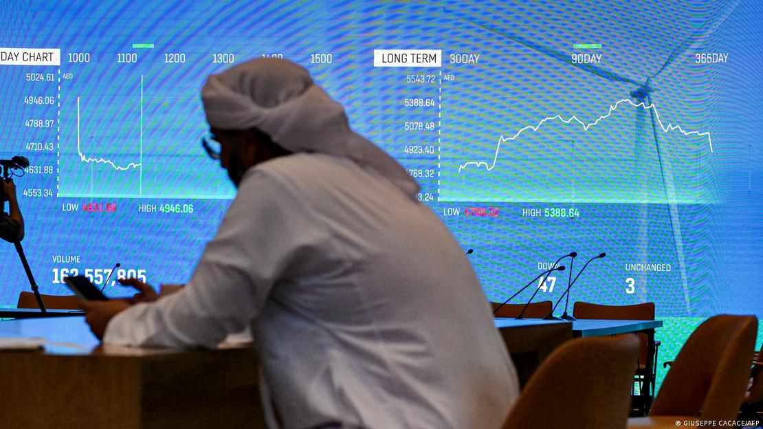 ایک شخص دبئی میں اسٹاک ایکسچینج میں نصب اسکرین کے قریب بیٹھ کر اپنے موبائل فون پر نظر ڈال رہا ہے، جہاں اسٹاک انڈیکسزدکھائی دے رہے ہیں 