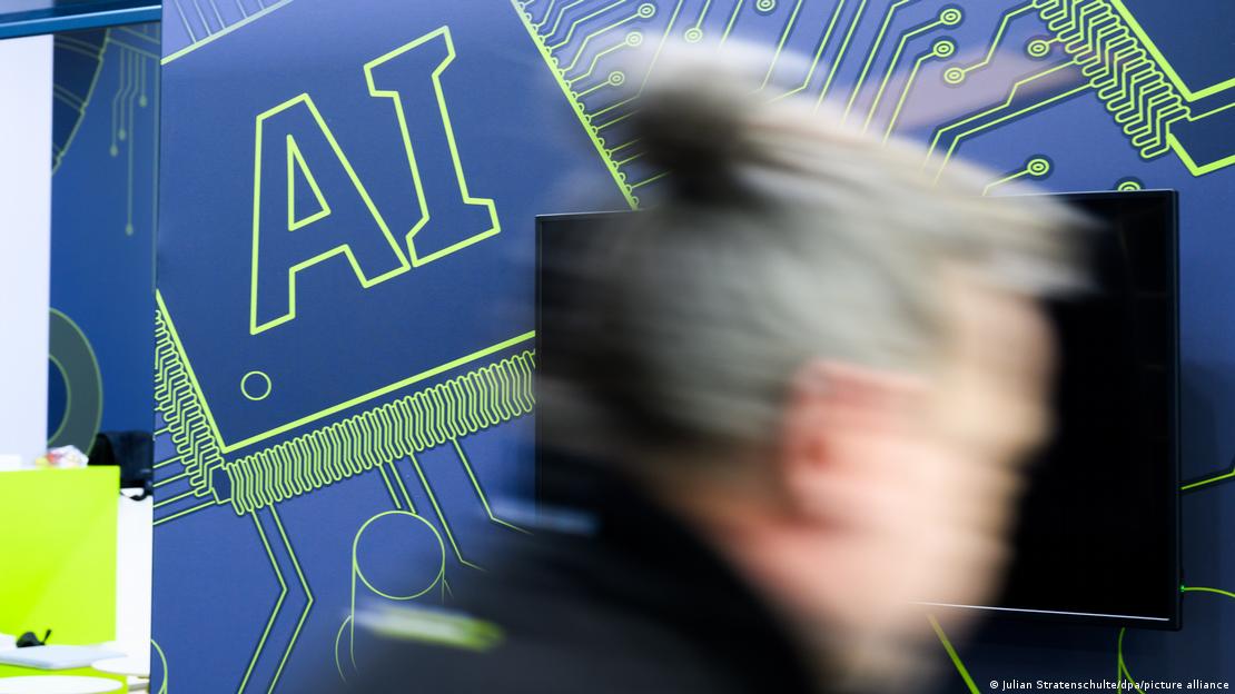 Un letrero que dice ‘AI’ se exhibe en la Feria Industrial de Hannover, 2025, y una persona pasa delante de él.