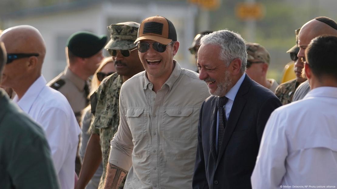 El secretario de Defensa de EE. UU., Pete Hegseth (con gorra de béisbol, gafas de sol y sonriente), camina junto a otro hombre sonriente con cabello canoso y traje. El secretario de Defensa de EE. UU., Pete Hegseth (con gorra de béisbol, gafas de sol y sonriente), camina junto a otro hombre sonriente con cabello canoso y traje.