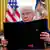 Donald Trump holds an executive order booklet in the Oval Office, with the American flag and Presidential standard in the background.